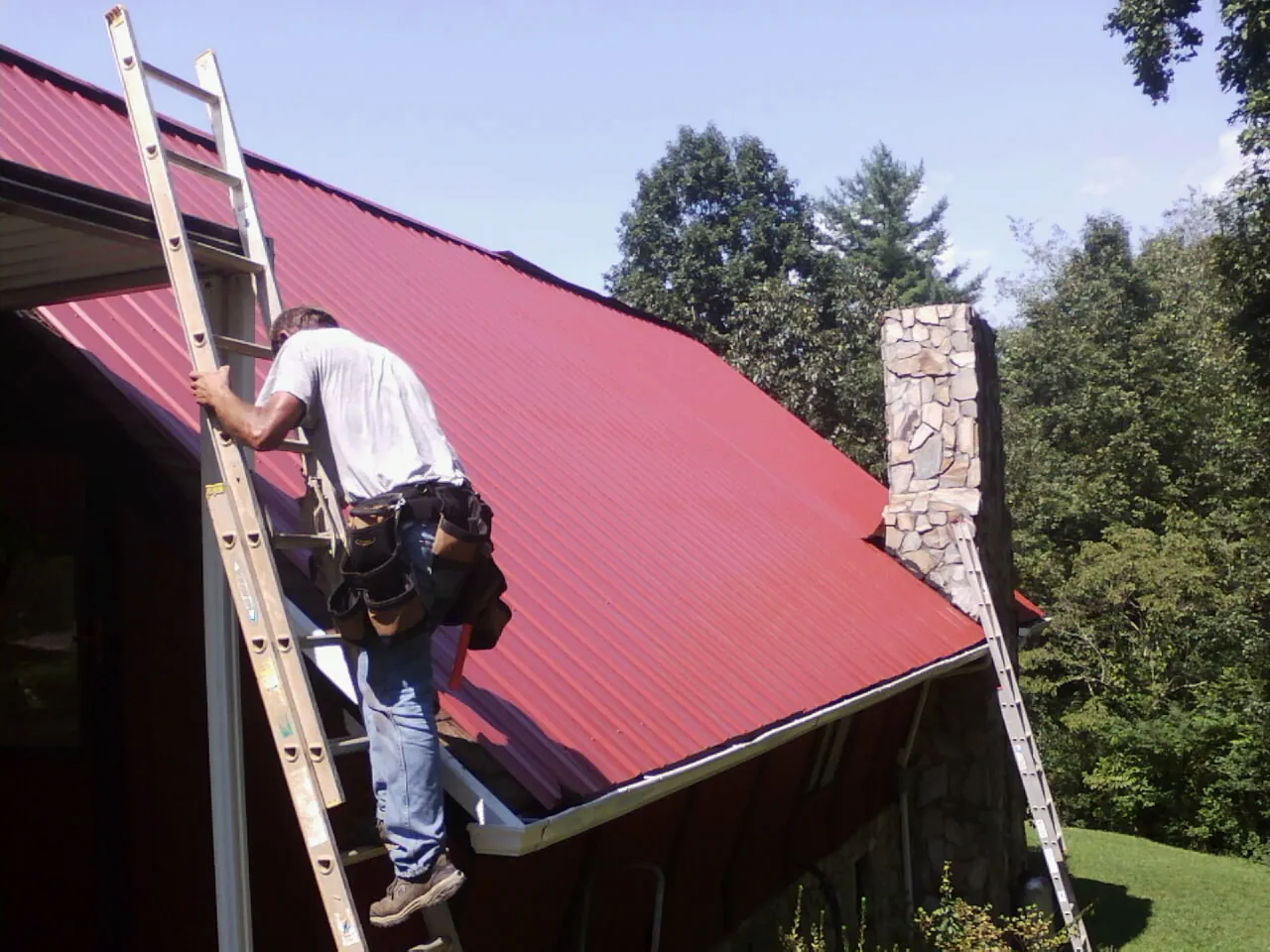 Roofer on a ladder beside a red metal roof and stone chimney — Soffit & Fascia Repair by Roofing Brothers in Thorndale
